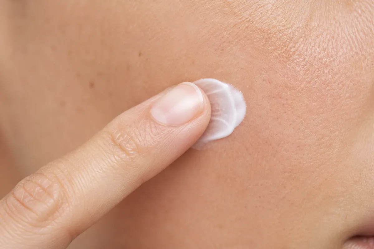 Main appliquant une crème hydratante sur une joue, gros plan texture peau, lumière douce, sans visage visible